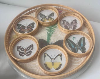 Peut inclure: Un ensemble de six dessous-de-verre ronds en bambou tissé avec un fond blanc et des motifs de papillons colorés. Les dessous-de-verre sont disposés en cercle sur un plus grand plateau en bambou tissé.