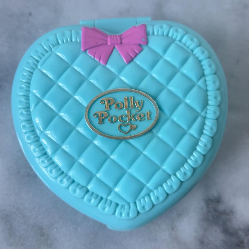 Pu&ograve; includere: Un giocattolo Polly Pocket a forma di cuore blu con un design trapuntato e un fiocco rosa. Il giocattolo ha un logo ovale dorato che recita "Polly Pocket" con un piccolo cuore.