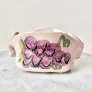 Puede incluir: Un pequeño plato de cerámica blanca con un diseño de uvas. Las uvas están pintadas en tonos de morado y verde.