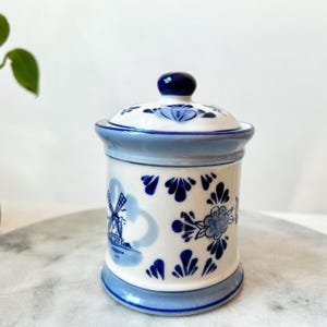 May include: A white ceramic canister with a lid, featuring blue hand-painted designs including a windmill, floral patterns, and decorative leaf motifs. The canister has a light blue rim at the base and top, with a dark blue knob on the lid.