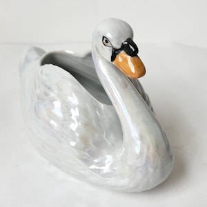 May include: A white ceramic swan planter with iridescent glaze. The swan has a long neck and a small head with a black eye and an orange beak.