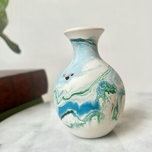 May include: A small ceramic vase with a marbled design. The vase has a white base, a light blue neck, and a body swirled with blue, green, and white. The vase has a rounded body and a narrow neck, suitable for displaying flowers.