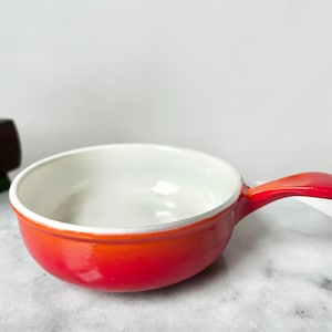 May include: A small, round, red and orange enameled cast iron pan with a white interior and a single handle. The pan is sitting on a marble surface. The pan is likely used for cooking and serving.