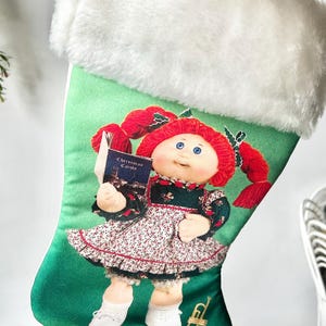 May include: A Christmas stocking with a green background featuring a doll illustration holding a book titled "Christmas Carols." The doll has red hair and is wearing a floral dress. The stocking has a white faux fur trim.