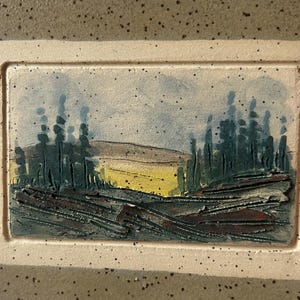 May include: A rectangular ceramic tile featuring a landscape painting. The artwork shows dark green trees, a yellow field, and a blue sky. The tile has a cream border and a textured surface.