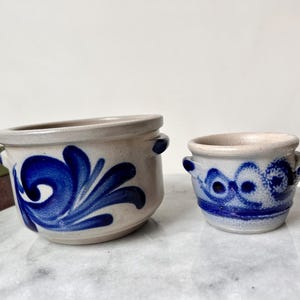 May include: Two small, beige ceramic pots with blue floral designs. The larger pot has a swirling pattern, while the smaller pot features a series of connected circles. Both pots have small handles.