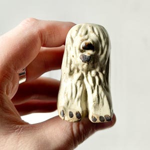 May include: A white ceramic figurine of a dog with a shaggy coat. The dog is sitting with its paws tucked under its body. The figurine has a brown nose and black eyes.