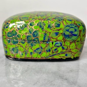 May include: A green and gold painted papier-mâché box with a floral design. The box is oval shaped and has a black base.