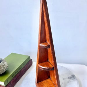 May include: A wooden corner shelf with three curved shelves. The shelf is a rich brown colour and has a triangular shape. A metal hook is at the top for hanging. The shelf is displayed on a white surface.