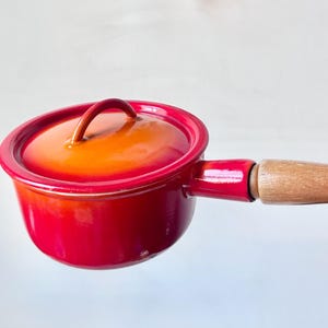 May include: A small, red enamelled saucepan with a matching lid. The lid and interior are orange. It has a wooden handle and is designed for cooking. The pot is likely made of cast iron or a similar material.