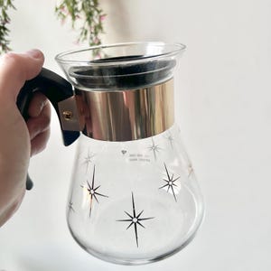 May include: A clear glass carafe with a black handle and a gold-coloured metal band. The carafe is decorated with black starburst designs. The carafe is likely vintage and suitable for serving beverages.