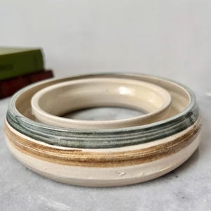 May include: A ceramic bowl set with a cream interior and a striped exterior in green, brown, and cream. The set includes a larger outer bowl and a smaller inner bowl, creating a nested design. The bowls are handmade.