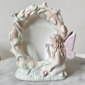 Op de afbeelding: Een keramische fotolijst met een knielende fee die een bloem vasthoudt. Het frame is omgeven door een boomachtig ontwerp met bladeren en een roze en wit kleurenschema. De fee heeft roze vleugels.