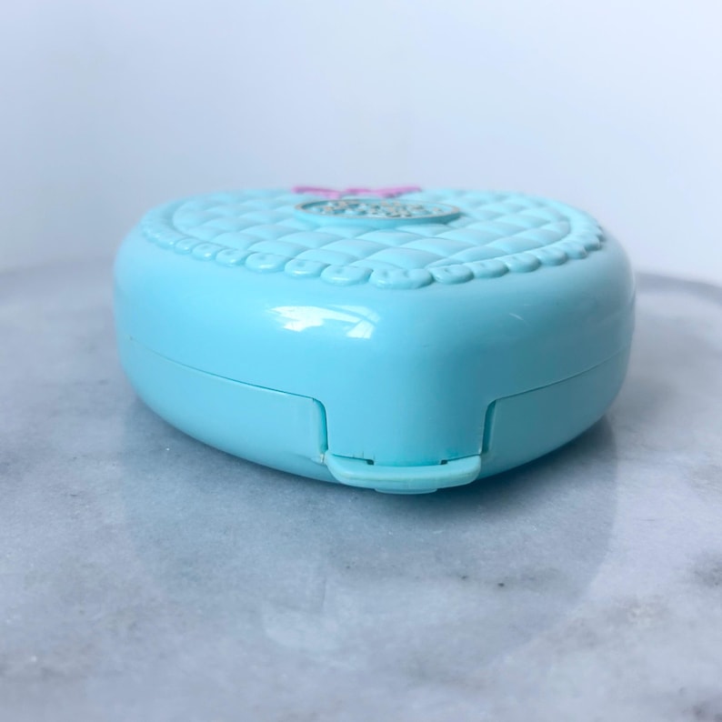 Pu&ograve; includere: Un compatto di plastica a forma di cuore di colore azzurro con un design trapuntato in rilievo e un fiore rosa e bianco sul coperchio.