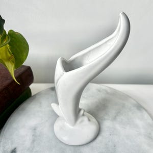 May include: A white ceramic vase with a unique, curved design. The vase has a rounded base and a tall, open top that curves upwards. The vase is smooth and glossy, with a minimalist aesthetic. It is a decorative item.