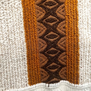 May include: A woven textile featuring a central vertical band of dark brown diamonds flanked by solid mustard yellow stripes and textured off-white panels. The bottom edge is finished with a row of white fringe, creating a decorative border.
