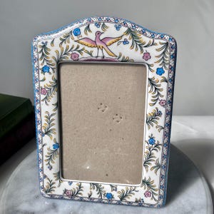 May include: A white ceramic picture frame with a colourful floral and bird design. The frame has a rectangular opening and is decorated with blue, pink, and yellow flowers and green leaves. A pink bird with yellow wings is at the top.