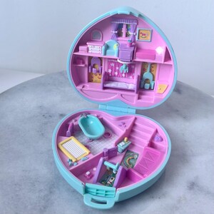 Pu&ograve; includere: Una casetta giocattolo in plastica blu e rosa a forma di cuore, aperta per rivelare due stanze. La stanza superiore ha un letto, una cassettiera e un cavallo a dondolo. La stanza inferiore ha una vasca da bagno, un box e uno scivolo.
