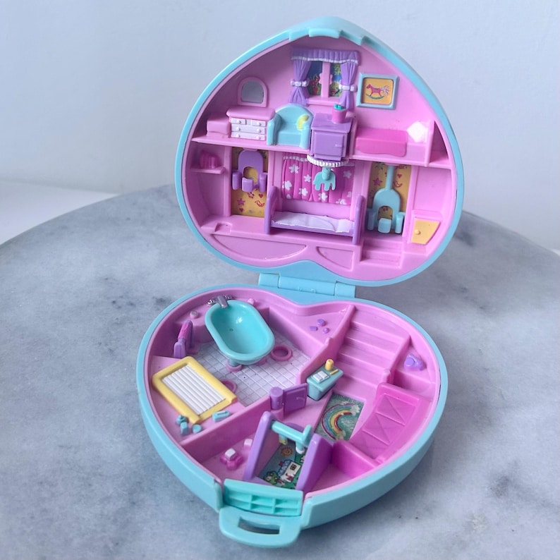 Pu&ograve; includere: Una casetta giocattolo in plastica blu e rosa a forma di cuore, aperta per rivelare due stanze. La stanza superiore ha un letto, una cassettiera e un cavallo a dondolo. La stanza inferiore ha una vasca da bagno, un box e uno scivolo.