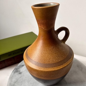 Può includere: Un vaso in ceramica marrone con manico, con fasce orizzontali di varie tonalità di marrone. Il vaso ha un collo stretto e un corpo arrotondato. Lo sfondo include un libro verde e una superficie di marmo.