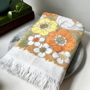 Op de afbeelding: Een witte rechthoekige handdoek met een herhalend bloemenpatroon in tinten oranje, geel en olijfgroen. De handdoek heeft een textuur van badstof en is afgewerkt met een witte franjerand langs de korte onderzijde van de handdoek.