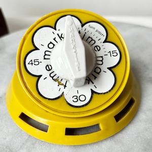 Op de afbeelding: Een felgele kookwekker met een witte bloemvormige wijzerplaat met zwarte cijfers 15, 30 en 45. Een witte centrale knop is gelabeld met Mark-Time en bevat de tekst turn past. De timer heeft een ronde basis met drie rechthoekige uitsparingen.