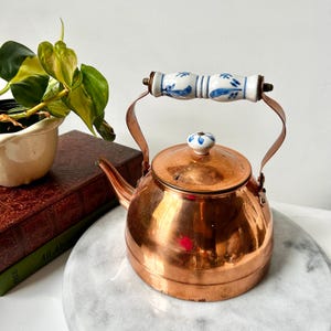 May include: A copper tea kettle with a ceramic handle and lid knob. The handle is white with blue floral designs. The kettle sits on a marble surface, with a plant and books in the background. The kettle is a vintage style.