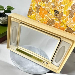 Puede incluir: Un espejo de tocador Lady Kenmore vintage de color crema con un espejo rectangular y dos luces laterales. El espejo está sobre una superficie similar al mármol gris, con una bolsa con estampado floral en el fondo.