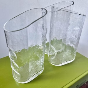 May include: Three clear, textured glass tumblers with an asymmetrical design. The glasses have a unique shape with a slanted top and a textured surface. They are arranged on a green surface.