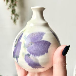 Może przedstawiać: Mały, kremowy ceramiczny wazon o zaokrąglonym korpusie i wąskiej szyi. Wazon jest ozdobiony dużymi, abstrakcyjnymi fioletowymi wzorami kwiatowymi. Wazon jest trzymany w dłoni, a tło jest rozmyte.