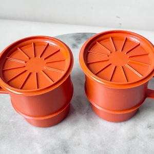 Puede incluir: Dos tazas de plástico naranja con tapas a juego. Cada taza tiene un asa y un diseño acanalado en la tapa. Las tazas están sobre una superficie gris claro.