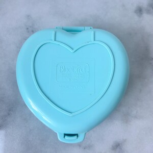Pu&ograve; includere: Una custodia a forma di cuore in plastica di colore azzurro chiaro con un logo Bluebird Toys e il testo "Made in China".