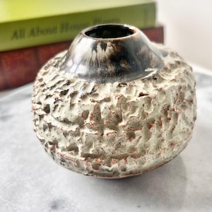 May include: A small, round ceramic vase with a textured, off-white exterior and a dark brown, glossy rim. The vase has a unique, organic shape and a rough, tactile surface, perfect for displaying small flowers or as a decorative accent.