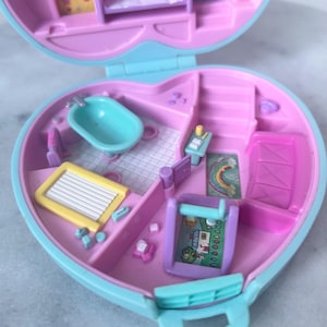 Pu&ograve; includere: Una casetta di giocattolo in plastica rosa e blu con una vasca da bagno, un letto, una scrivania, un tappeto arcobaleno e una lavagna. La casa &egrave; aperta e l'interno &egrave; visibile.