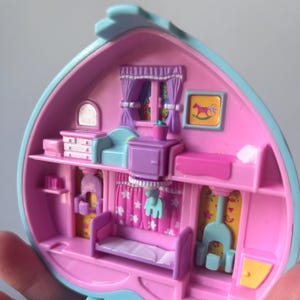 Pu&ograve; includere: Una casetta di giocattolo in plastica rosa e blu con un letto, una cassettiera, un cavallo a dondolo e un camino. La casa &egrave; aperta e l'interno &egrave; visibile.