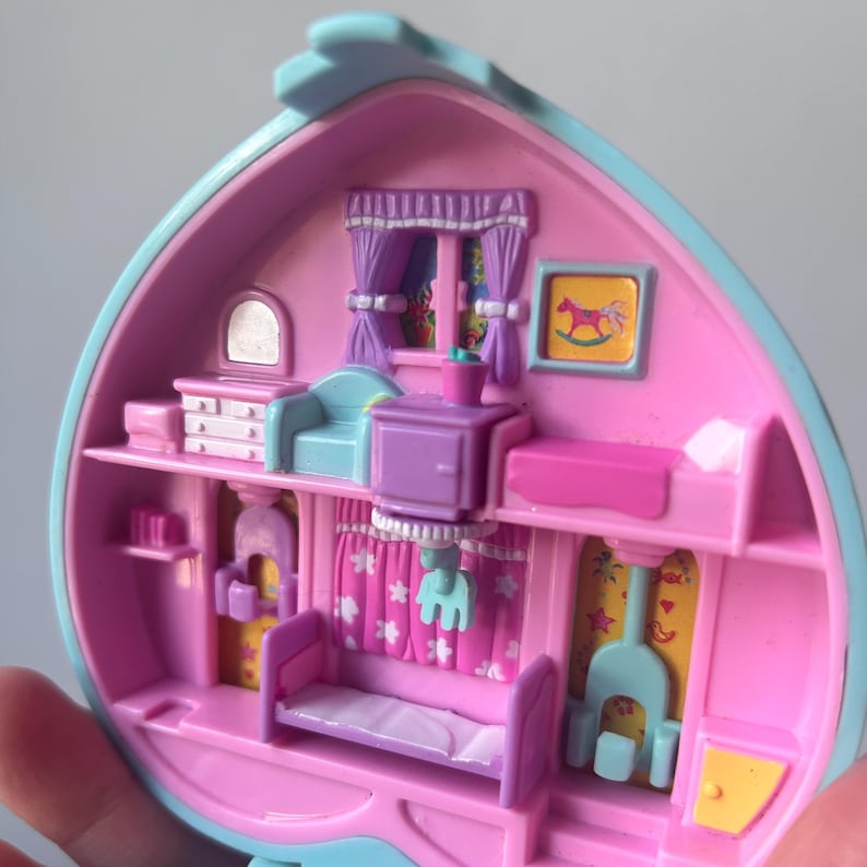 Pu&ograve; includere: Una casetta di giocattolo in plastica rosa e blu con un letto, una cassettiera, un cavallo a dondolo e un camino. La casa &egrave; aperta e l'interno &egrave; visibile.