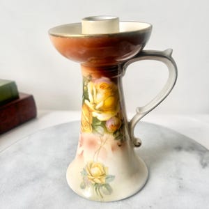 May include: A vintage ceramic candlestick holder with a handle. The holder features a cream base with a floral design of yellow roses and green leaves. The top is a deep orange color with a cream-colored candle cup.
