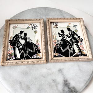 Può includere: Due immagini incorniciate di silhouette di una coppia in un contesto nuziale. Le silhouette sono nere su uno sfondo bianco con accenti floreali. Le cornici sono in legno chiaro.
