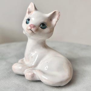 May include: A white ceramic cat figurine with blue eyes and a pink nose and ears. The cat is in a seated position, with its paws tucked in. The figurine has a glossy finish and is a decorative item.