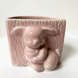 Könnte beinhalten: Ein rosafarbener Keramik-Pflanztopf in Form eines Kaninchens. Der Topf hat eine strukturierte Oberfläche und eine weiße Glasur.