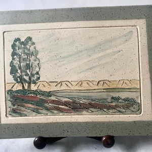 Peut inclure: Pièce d'art en céramique rectangulaire représentant un paysage. L'œuvre présente un arbre, de l'eau et des montagnes, le tout dans des tons sourds de vert, de brun et de beige. La pièce est encadrée d'une bordure grise mouchetée. Mesures en centimètres.
