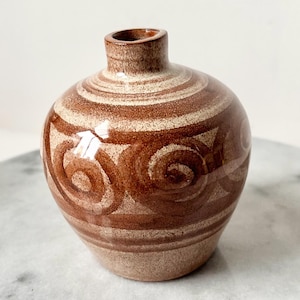 May include: A small, round ceramic vase with a narrow neck. The vase is primarily brown with a spiral design in a lighter tan color. The design features concentric circles and swirled patterns, creating a decorative effect. The vase has a smooth, glossy finish.