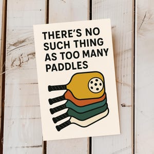 May include: A cream-coloured print with the text "THERE'S NO SUCH THING AS TOO MANY PADDLES." Below the text are stacked pickleball paddles in yellow, orange, and green with black handles. The paddles are on a white wooden background.