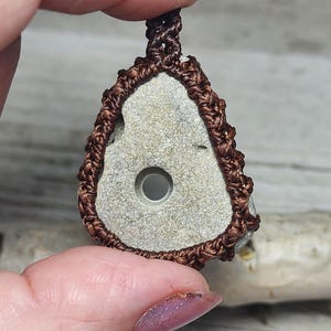 May include: A handcrafted pendant featuring a light gray stone with a central hole, encased in a brown macrame design. The pendant is suspended from a matching macrame cord, showcasing a natural, earthy aesthetic.