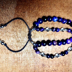 May include: Two beaded bracelets with dark blue and clear beads. The bracelets are connected by a black cord with silver-colored metal beads at the ends. The bracelets are on a wooden surface.