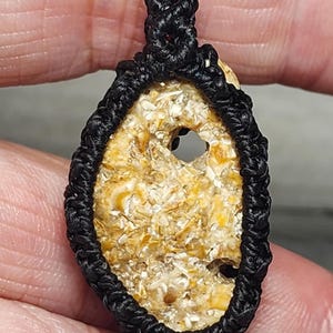 May include: Oval-shaped pendant with a textured, beige and yellow stone center, framed by a black woven cord. The stone has a hole in the center. The pendant is held in a hand.