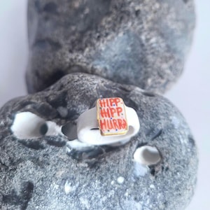 May include: A white ceramic ring with a red and gold rectangular inlay. The inlay has the words "Hipp Hipp Hurra" written on it.
