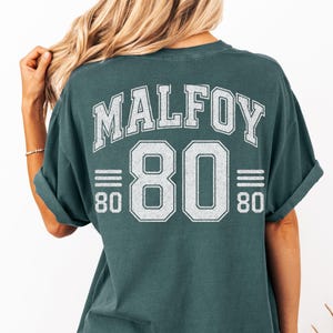 May include: A teal-colored t-shirt with the word "MALFOY" arched across the upper back in white block letters. Below, the number "80" is centered, also in white, with horizontal lines above and below, and the number "80" on each side.