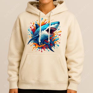 K&ouml;nnte beinhalten: Ein cremefarbener Kapuzenpullover mit einer auff&auml;lligen Grafik eines blauen Hais mit offenem Maul, umgeben von farbenfrohen Spritzern in Rot, Orange, Gelb und Blau. Der Kapuzenpullover hat eine Kapuze mit Kordelzug und eine Vordertasche.