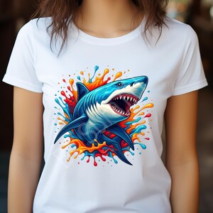 K&ouml;nnte beinhalten: Wei&szlig;es T-Shirt mit einem auff&auml;lligen Grafikdesign eines blauen Hais mit offenem Maul, umgeben von farbenfrohen Spritzern in Rot, Orange und Blau. Das Haidesign ist detailliert und lebendig, geeignet f&uuml;r den t&auml;glichen Gebrauch.